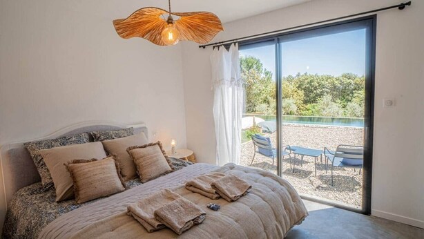 Une chambre dotée d’un grand lit, d’un luminaire suspendu et offrant une vue sur l’espace extérieur avec piscine.