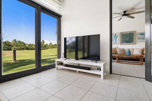 Una sala de estar moderna con un televisor de pantalla plana grande sobre un pedestal blanco, un ventilador de techo y vistas a un césped verde a través de puertas de vidrio amplias.
