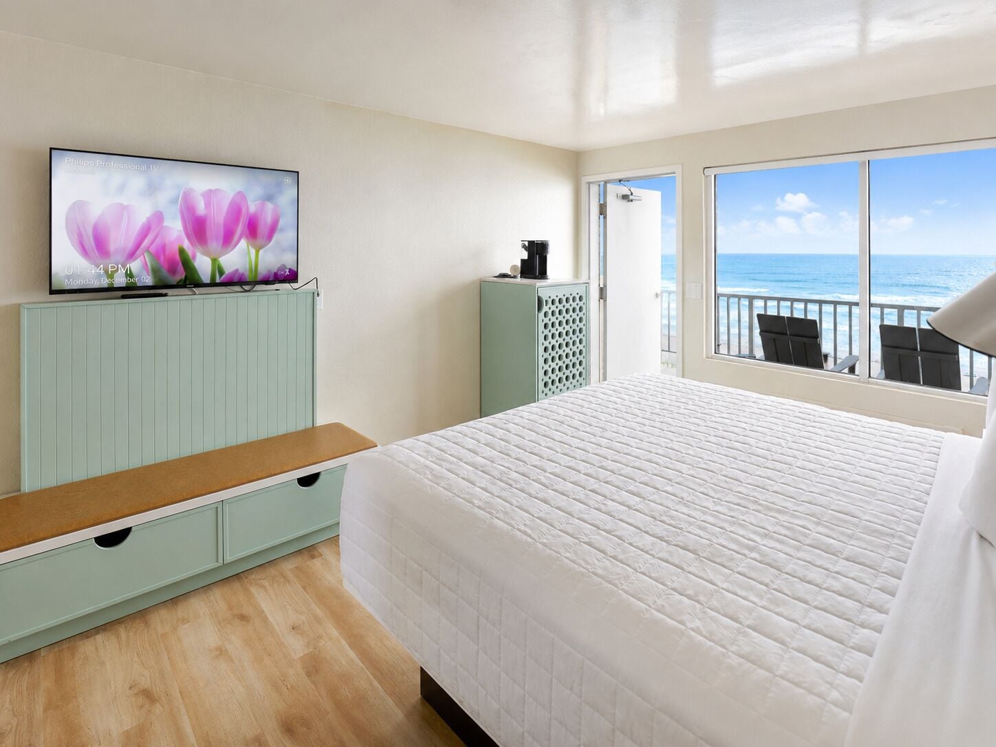 A hotel room with a bed, a television on a stand, a view of the ocean through a balcony, and a wooden floor.
