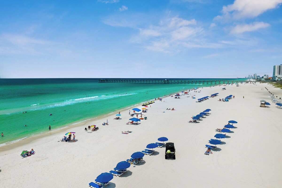 A beach with rows of blue umbrellas, a pier in the background, and a clear sky.