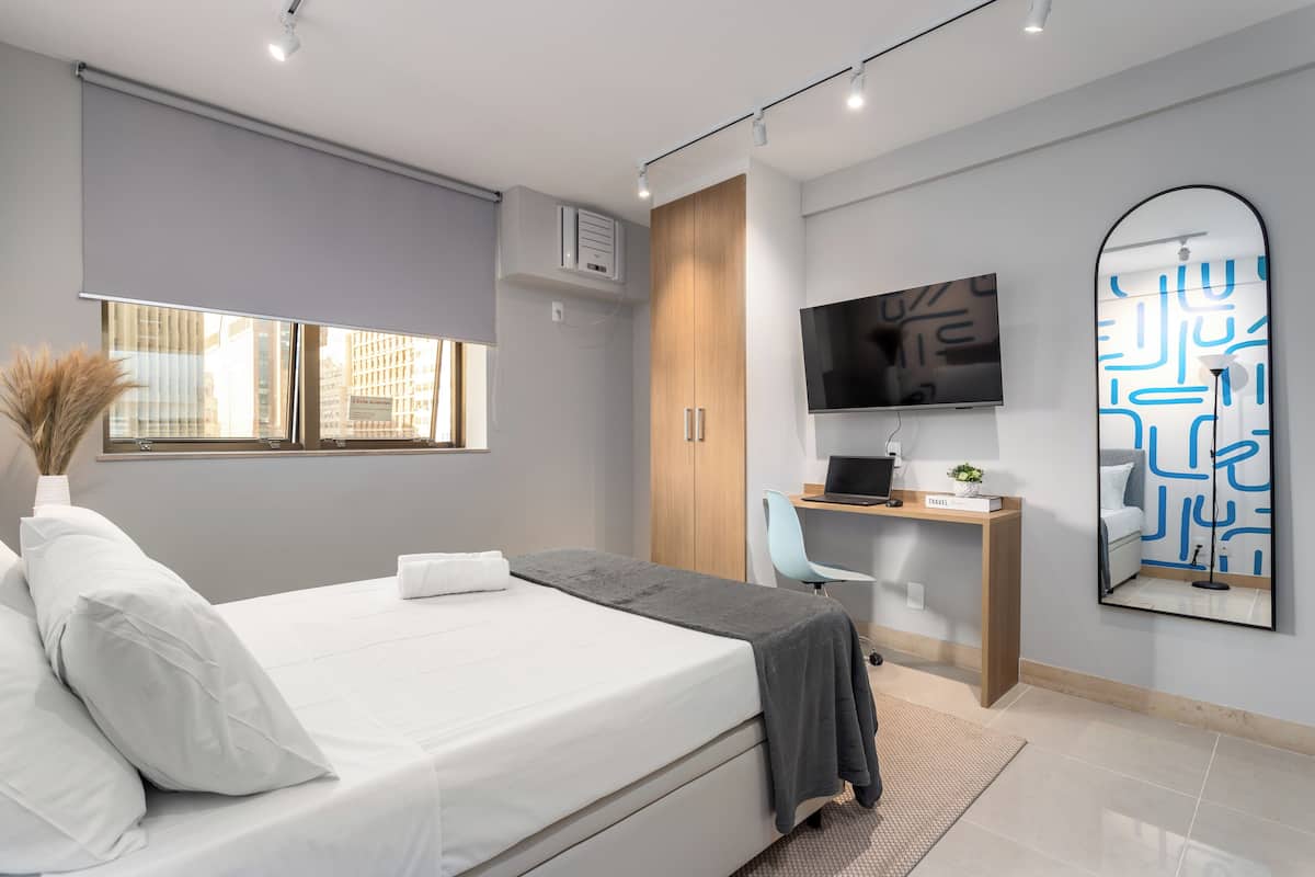 Quarto moderno com cama, escrivaninha, televisão e uma janela grande.