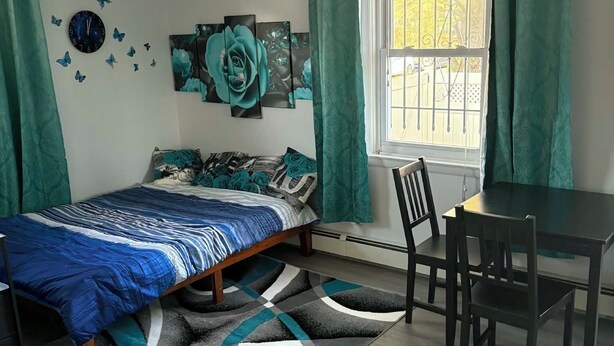 Un dormitorio con cama, un escritorio y una silla. Hay una ventana con cortinas, un reloj y decoración en la pared.