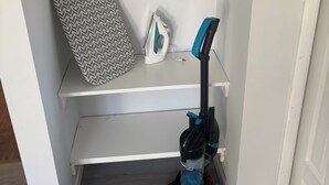 Un rincón de una habitación con una pared blanca, un estante con una tabla de planchar y un planchador, y una aspiradora en el suelo.