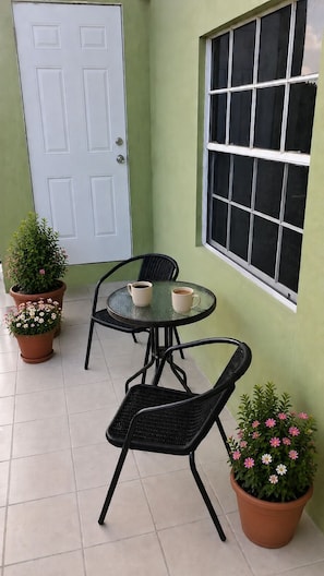 Een klein terras met een ronde tafel en twee stoelen, een witte deur en een raam met uitzicht.