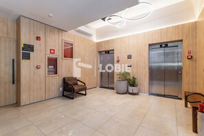 Um hall moderno com painéis de madeira, dois elevadores, um balcão de recepção e uma área de espera com cadeiras.