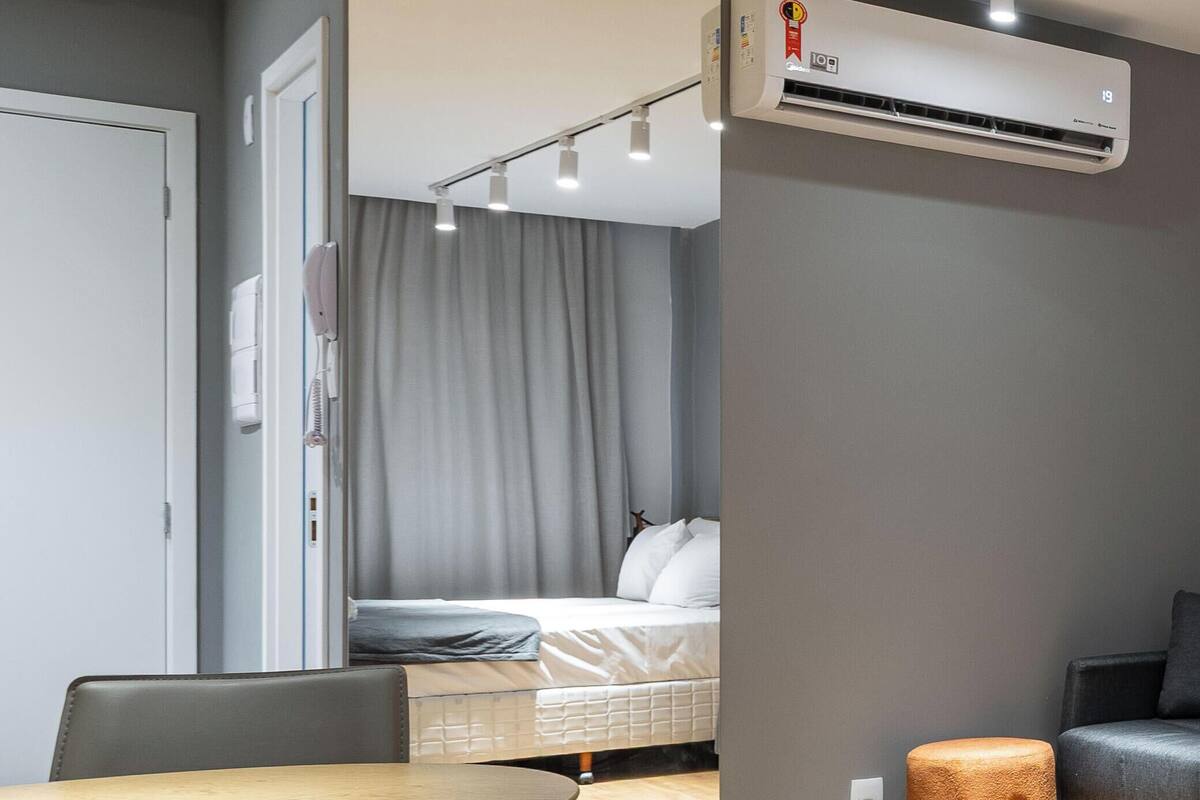 A modern hotel room with a wooden dining table, a small stool, a bed with a gray curtain, and an air conditioning unit.