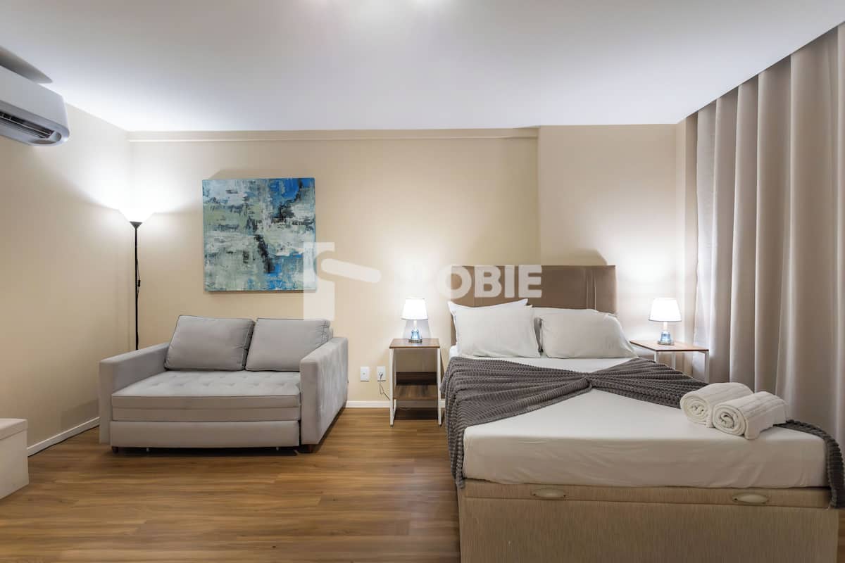 A hotel room with a bed, a sofa, a nightstand with lamps, a painting on the wall, and an air conditioning unit.