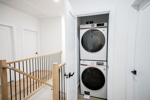 A laundry room with two stacked front-loading washing machines and a wooden staircase with black railings. - 3-bedroom townhome in welcoming Dallas with AC for comfort (Dallas)