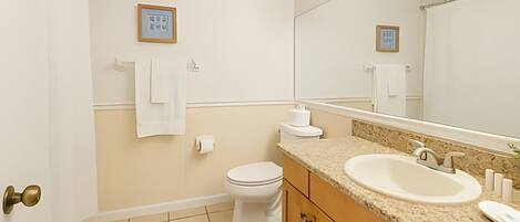 A bathroom with a toilet, sink, and mirror. There are towels hanging on the wall and a picture frame.