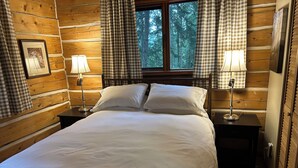A log cabin bedroom with a large bed, two bedside tables with lamps, checkered curtains, and a window with a forest view. - Welcome to Cloud 99! Cabin has privacy, quiet, awesome view! Recently updated. (Mayne Island)