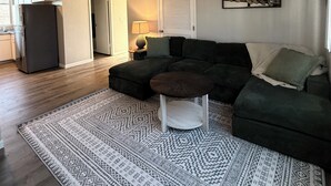 A living room with a dark green sofa, a patterned rug, and a small table. A kitchen with a refrigerator and cabinets is visible in the background. - Workman’s Rest Pt. 2 – Quiet Upstairs 2BR for Working Travelers (Gillette)