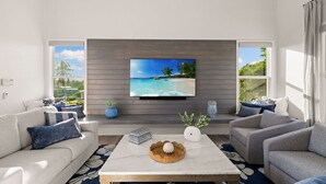 Ein modernes Wohnzimmer mit einem großen Flachbildfernseher an einer Holzwand, zwei Sofas mit gemusterten Kissen, einem Marmortisch und Blick auf einen tropischen Strand durch große Fenster.