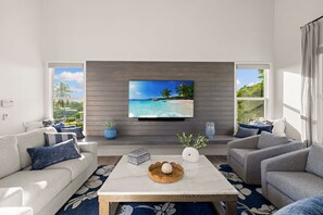 A modern living room with a large flat-screen TV mounted on a wooden wall, two sofas with patterned cushions, a marble coffee table, and a view of a tropical beach through large windows. - Twin Tides - Bayfront Views + Steps to Bonita Beach (Bonita Springs)