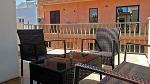 Un balcon extérieur aménagé avec des meubles en osier et une table en verre, offrant une vue sur un immeuble aux murs et balcons orange.