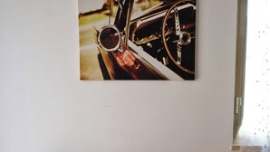 Un coin repas avec une table en bois, des chaises blanches et un tableau accroché au mur représentant l'intérieur d'une voiture.