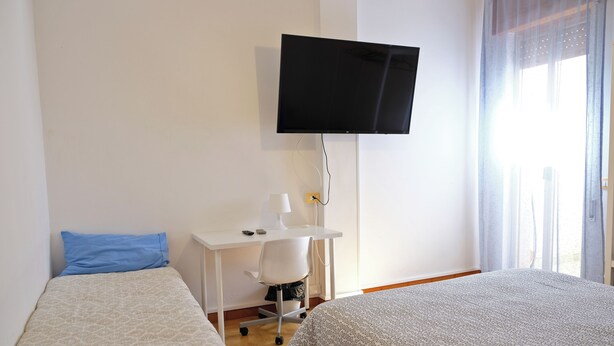 Une chambre d’hôtel avec deux lits, un bureau avec une chaise, un téléviseur fixé au mur et une fenêtre avec des rideaux.