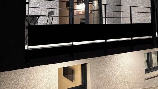 Un bâtiment moderne doté d’un balcon, d’une cuisine visible à travers une porte et d’un mur à la texture particulière.