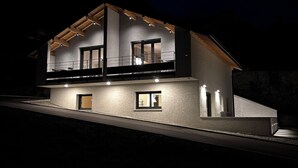 Une maison moderne à deux étages, dotée d’un balcon, de grandes fen êtres et d’un éclairage extérieur soigné pour la nuit.