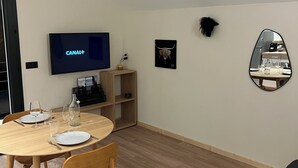 A modern living room with a round wooden dining table, two chairs, a flat-screen TV, a small shelf with decorative items, a framed picture, and a mirror on the wall.