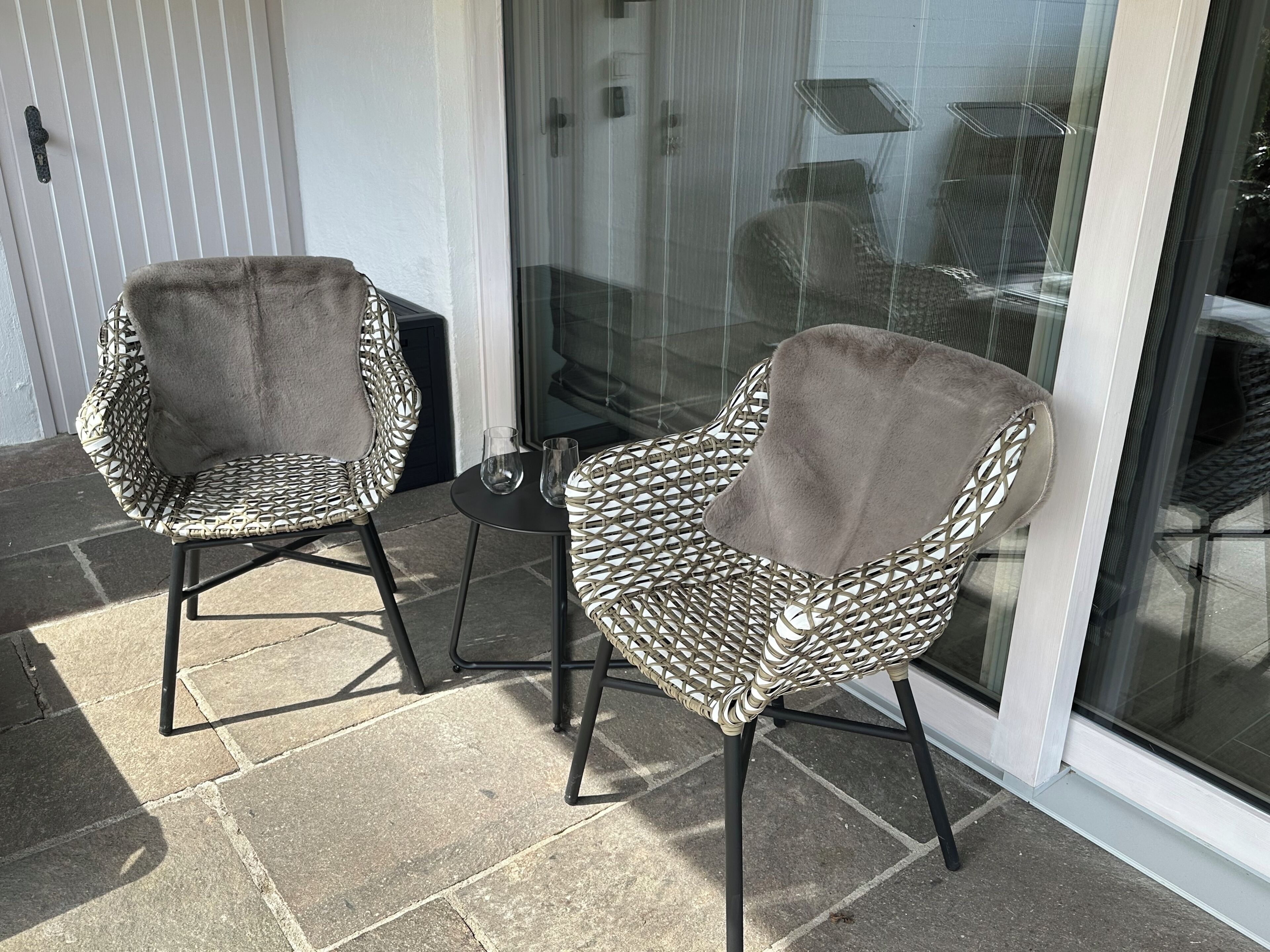 Dos sillas de mimbre con asientos y respaldos acolchados, ubicadas en un patio con puerta de cristal y una mesita con dos vasos.