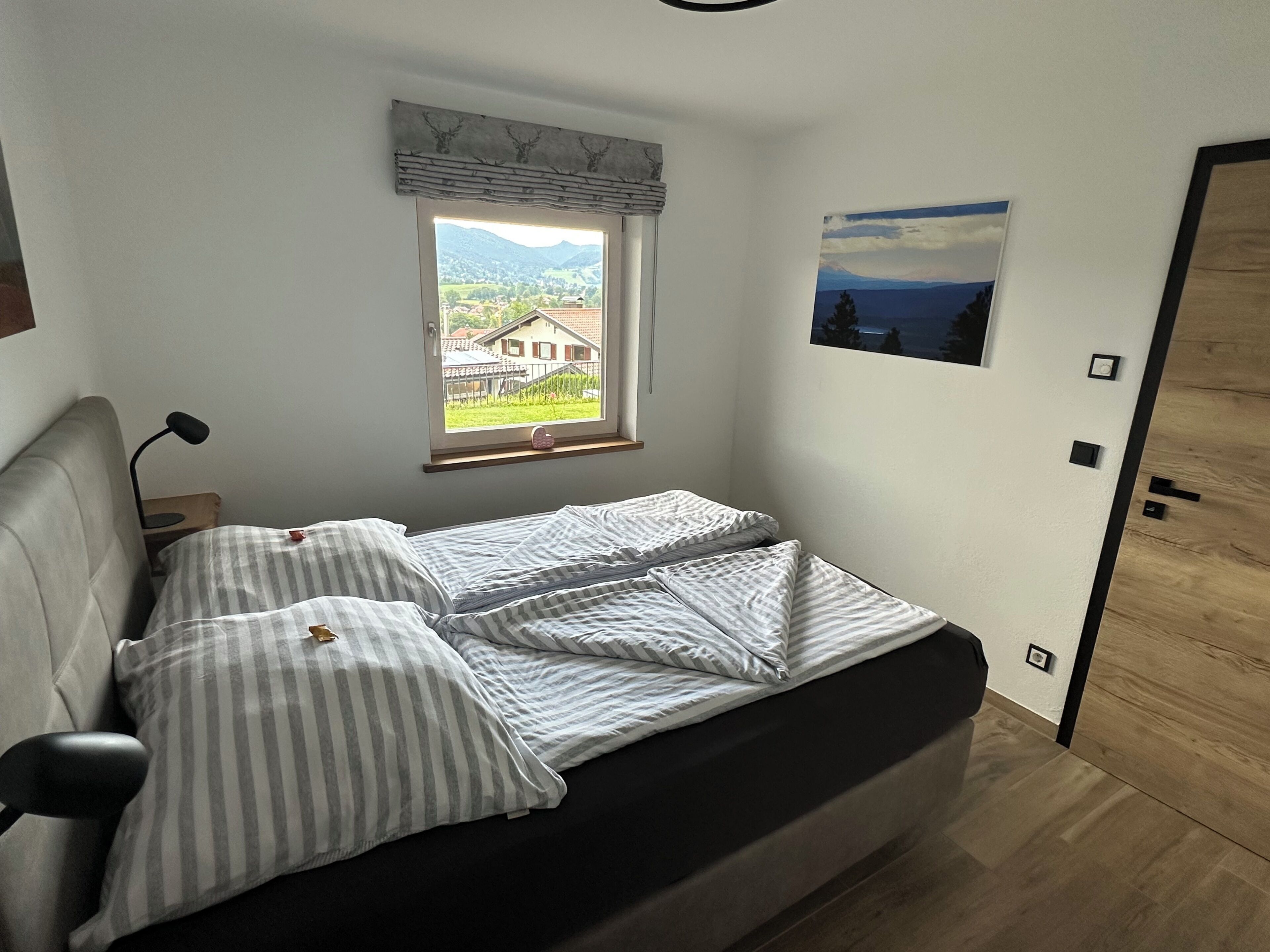 Ein Schlafzimmer mit Bett, Fenster mit Blick auf ein Haus und Berge, einer Tür und einem Wandbild.