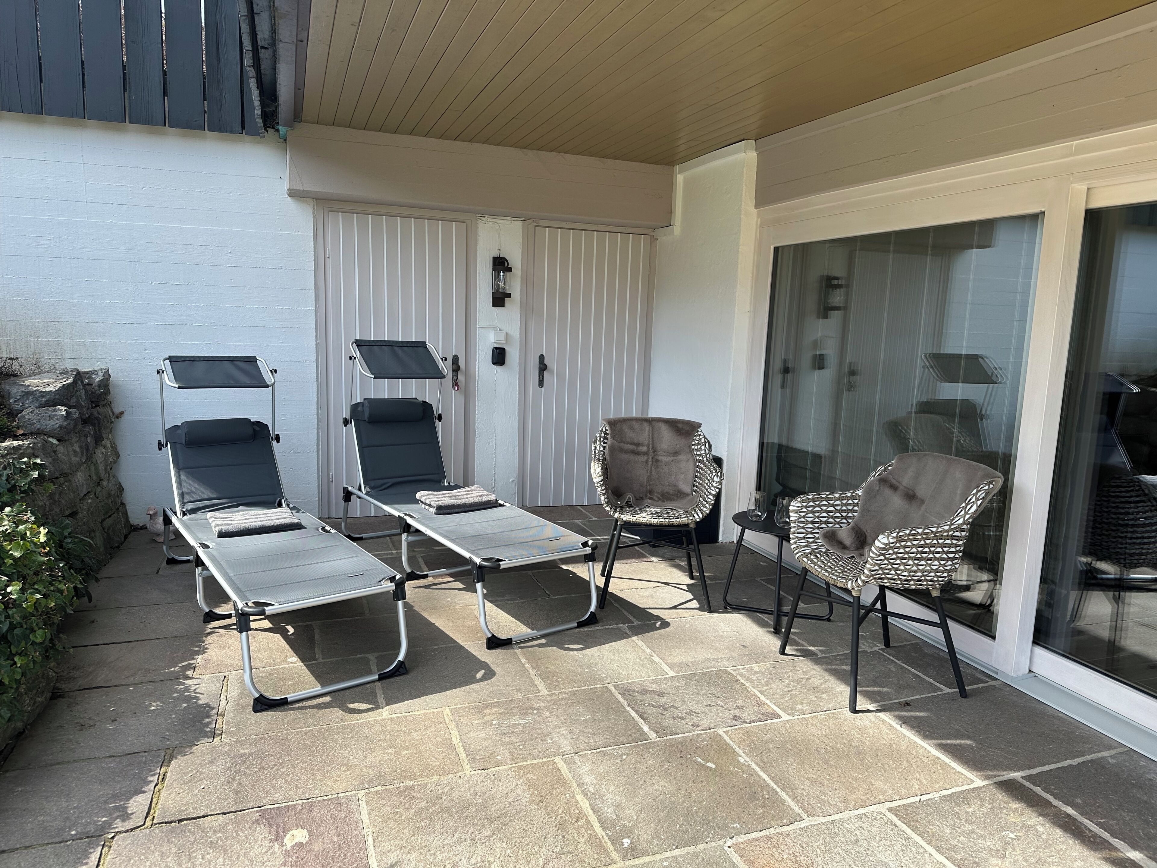 A patio with two lounge chairs, a cushioned chair, and a glass door leading to a covered area.