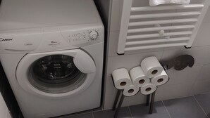En tvättmaskin med frontmatning, ett kontrollpanel, en handduksställning med ett hopvikt handduk och en hög toalettpapper på en liten pall.