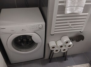 A front-loading washing machine with a control panel, a towel rack with a folded towel, and a stack of toilet paper on a small stool. - Villa Nižić Makarska -Two Bedroom Apartment With Terrace And Hot Tub (Lux F) (Makarska)