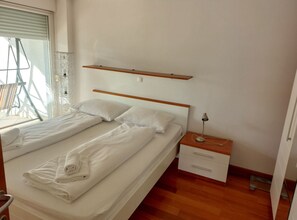 A bedroom with a bed, a bedside table, a lamp, and a window with blinds. - Villa Nižić Makarska - One Bedroom Apartment With Balcony (Marina) (Makarska)