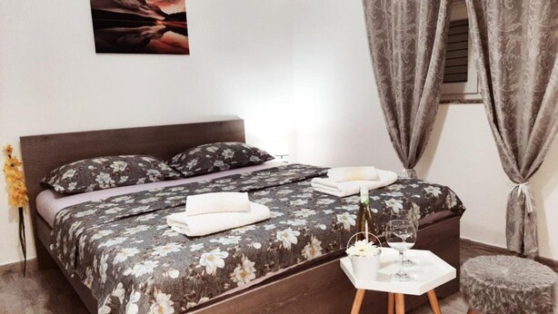 A bedroom with a bed, a bedside table with a bottle and glasses, a small stool, and a window with curtains. - Apartments Tomislav - Two Bedroom Apartment with Terrace and Sea View (Vrsi)