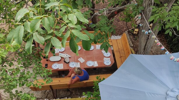 Ein Essbereich im Freien mit einem Holztisch für zwei Personen, umgeben von Grünpflanzen und einem blauen Sonnenschirm.