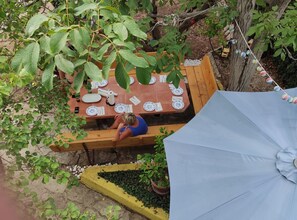 Un espace de restauration en plein air avec une table en bois dressée pour deux personnes, entourée de verdure et surplombée d’un parasol bleu.