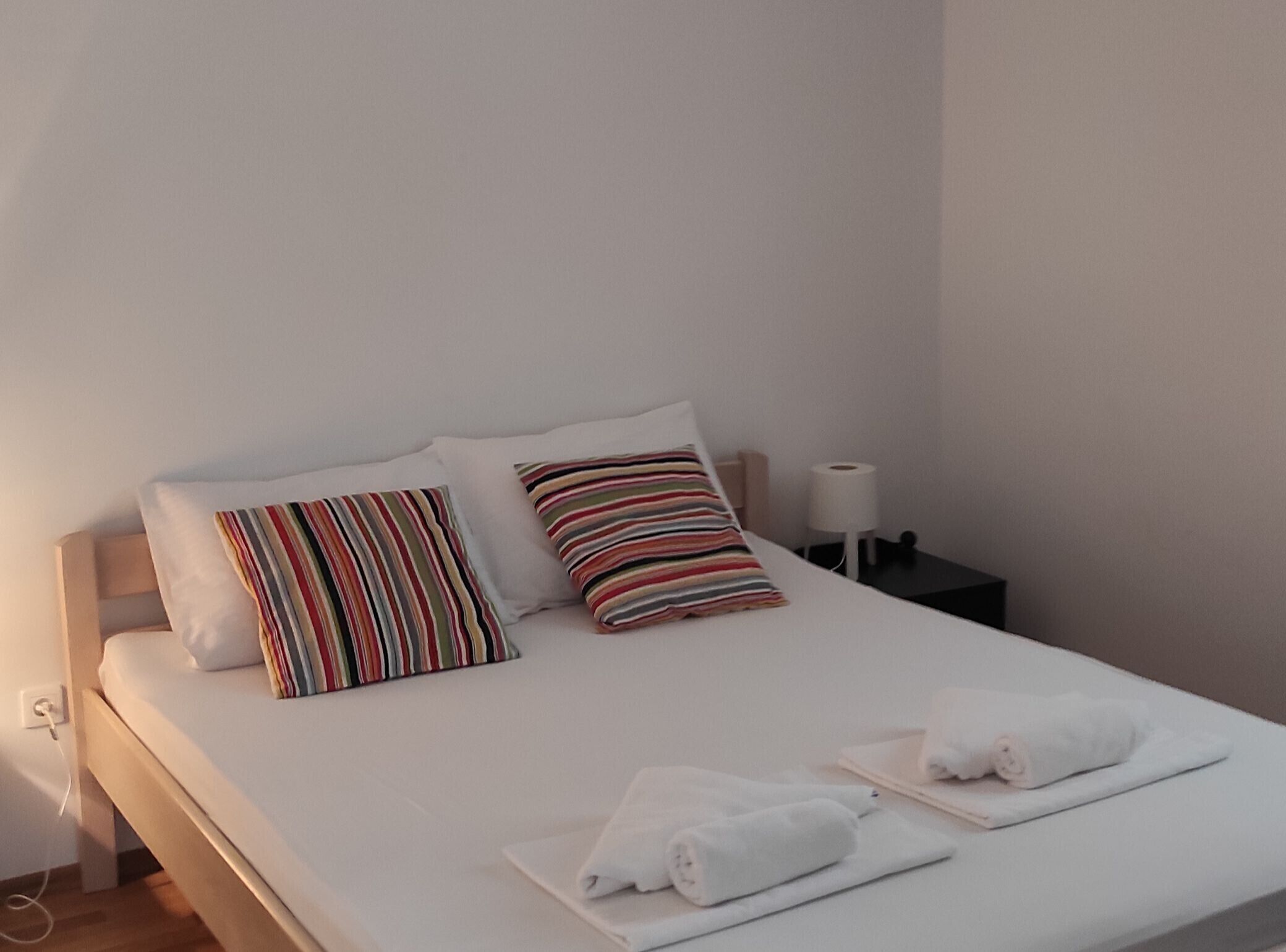 En pænt redt seng med hvide sengetøj og stribede puder, et natbord med en lampe, og sammenrullet håndklæder på sengen.