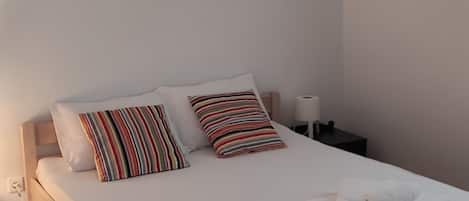 A neatly made bed with white linens and striped pillows, a bedside table with a lamp, and rolled towels on the bed.