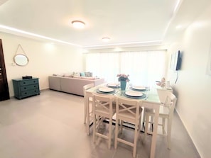 A dining area with a round table set for four, white chairs, a sofa, a small cabinet, and a TV mounted on the wall. - Punta Cana | 3 Min to Bibijagua Beach | 3 Pools (Punta Cana)