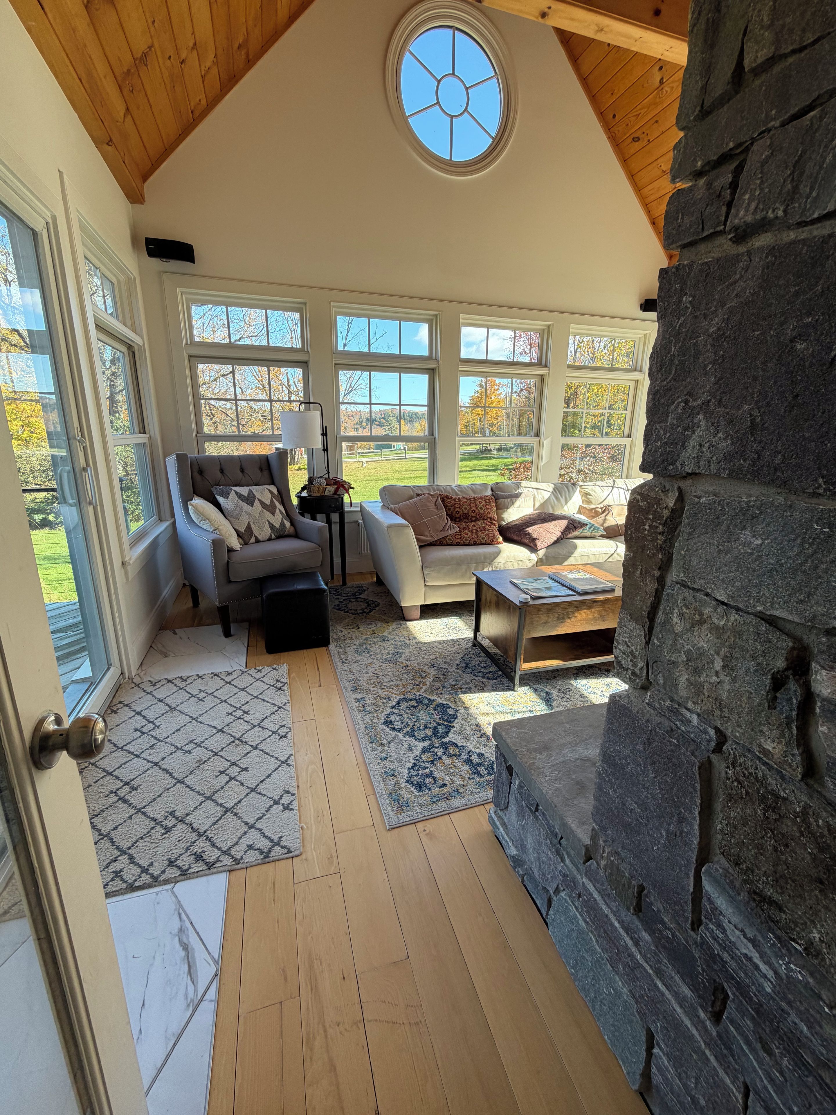 A cozy living room with a stone fireplace, large windows, and a wooden ceiling.