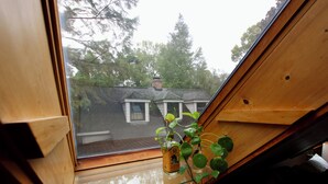 Une lucarne dotée d’un cadre en bois et d’une vitre, à travers laquelle on peut apercevoir une maison et des arbres.