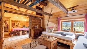 A rustic wooden cabin interior with a sofa, a wooden chest, a bench, and a window with a view of the outdoors.