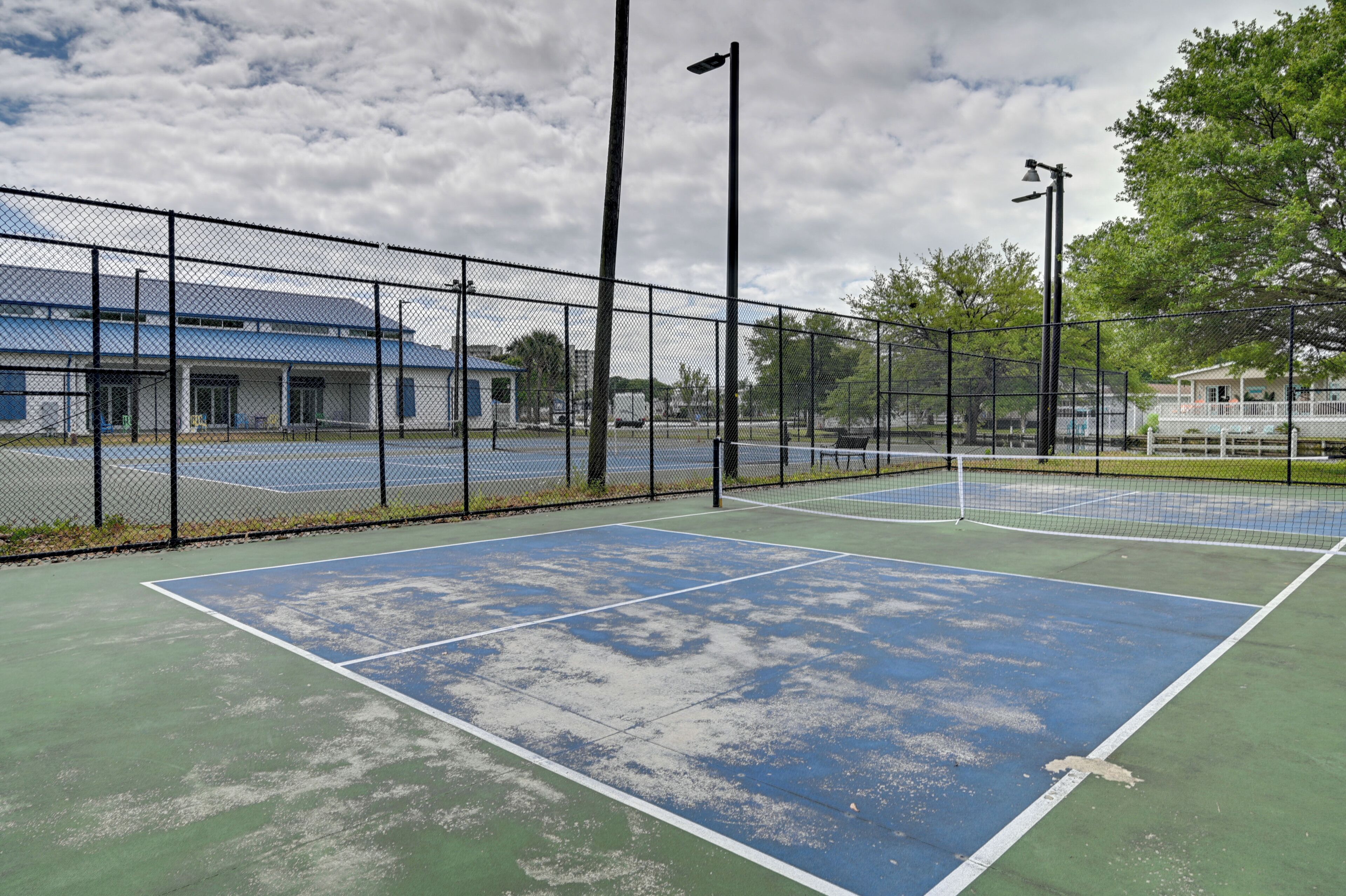 Un court de tennis avec un filet, entouré d’une clôture en treillis métallique, et un bâtiment avec un toit bleu en arrière-plan.
