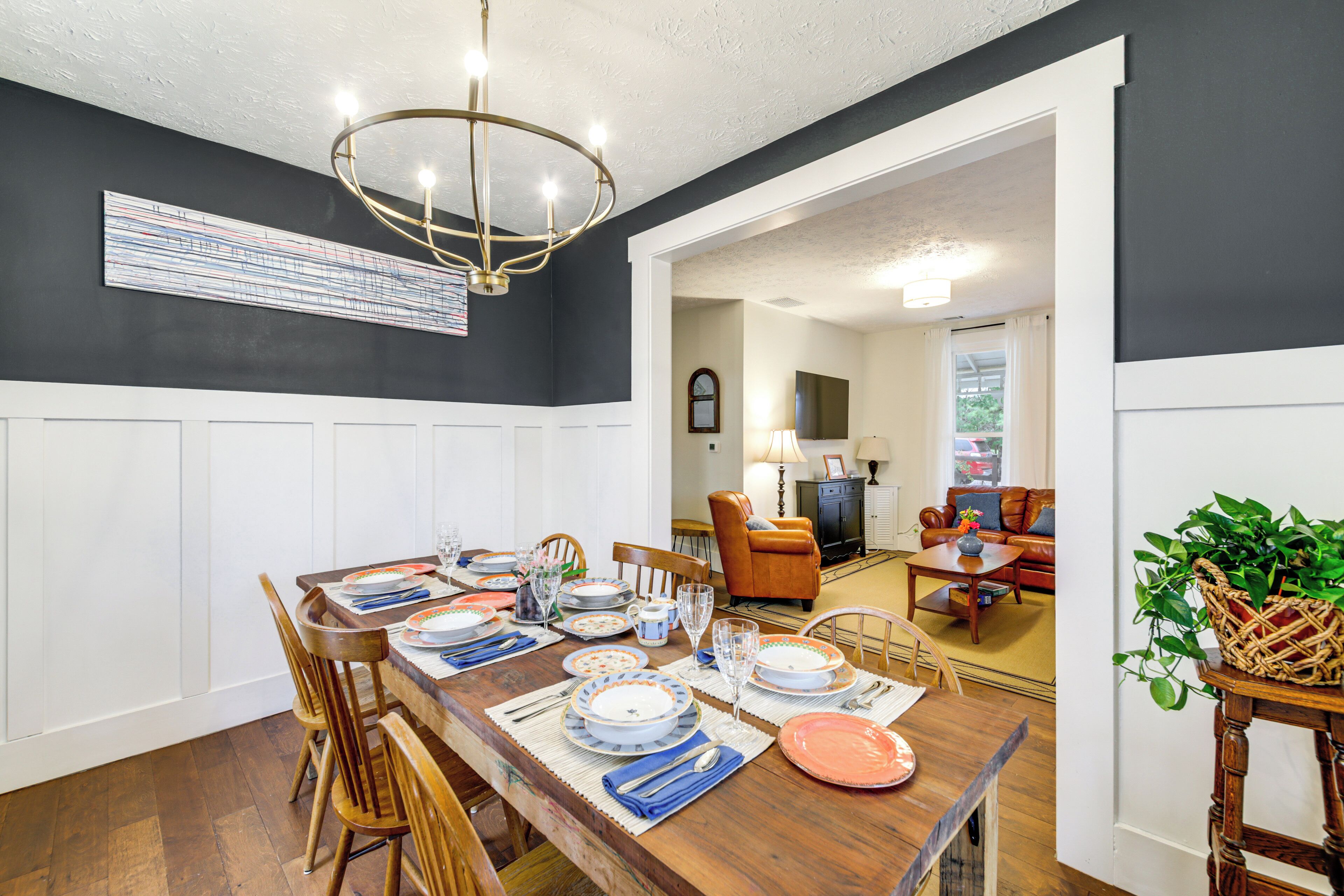 A dining area with a wooden table set for a meal, flanked by wooden chairs. A chandelier hangs from the ceiling, and a large abstract painting is on the wall.