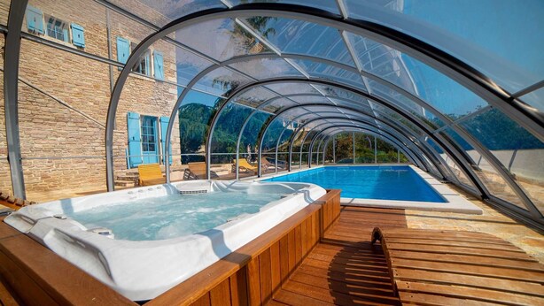 Una piscina cubierta al aire libre con hidromasaje, terraza de madera y edificio de piedra con contraventanas azules.