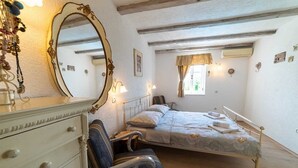 Un dormitorio con cama, una silla, tocador, espejo y ventana con cortinas.