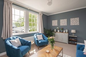 Un salón con dos sillones azules, una mesa de centro de madera, un radiador blanco y una ventana con cortinas.