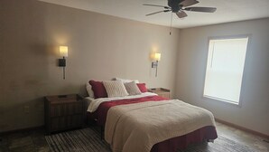 Une chambre à coucher comprenant un lit, deux tables de chevet, un ventilateur de plafond, une fenêtre avec stores et deux lampes fixées au mur.