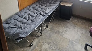 Une petite pièce avec une porte en bois, une fenêtre avec des stores, un matelas gris posé sur un cadre en métal et une petite poubelle.