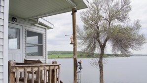 A wooden deck with a railing, a white building with windows, a tree, and a boat by the water. - Spacious Lakeside house on Long Lake Saint Agatha! (Saint Agatha)
