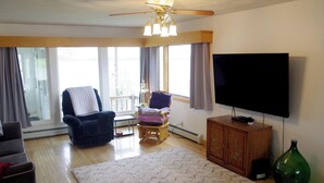 A living room with a ceiling fan, a flat-screen TV, a wooden cabinet, and a large window with curtains. - Spacious Lakeside house on Long Lake Saint Agatha! (Saint Agatha)