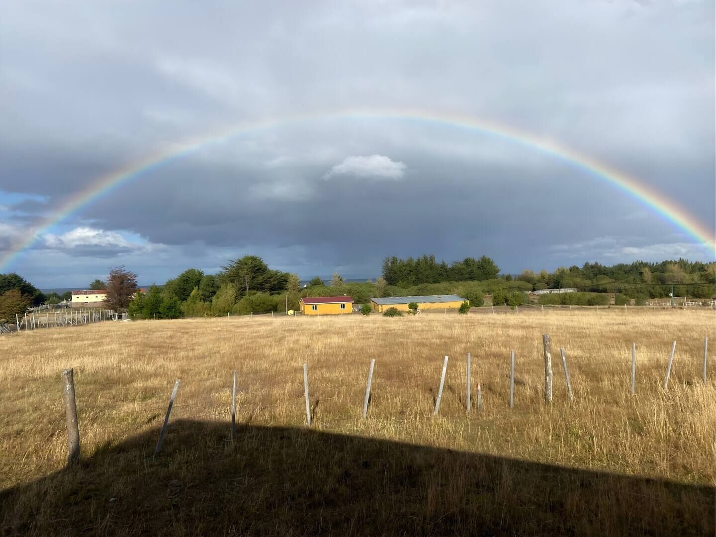 Un paysage rural avec un arc-en-ciel, un bâtiment jaune et un champ clôturé.