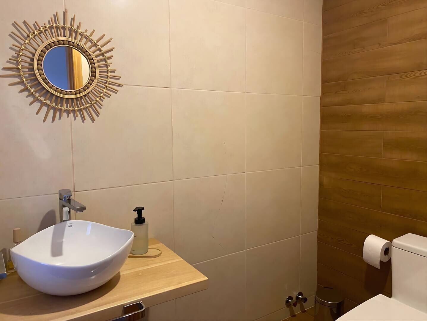 A bathroom with a white sink, a mirror with a sunburst frame, a soap dispenser, and a toilet.