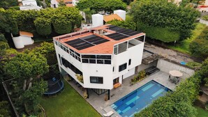 A modern two-story house with a rooftop solar panel array, a swimming pool, and a well-maintained garden.